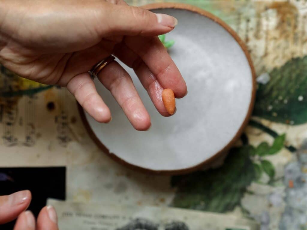 A hand is holding a small, oval-shaped bit or orange marzipan, between the thumb and index finger. In the background, there is a white bowl on a table with floral patterns and sheet music.